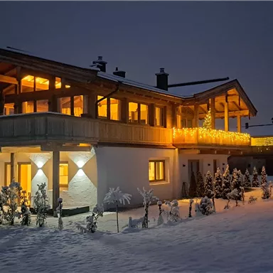 A cozy house in the snow, illuminated by warm light. In the background, additional buildings and snow-covered trees can be seen.