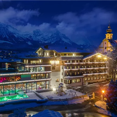 A lit hotel in a winter landscape at night. In the background, snow-covered mountains and a church can be seen.