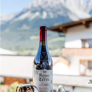 A bottle of red wine is on a table with a wine glass next to it. In the background, the mountains and a blue sky can be seen.