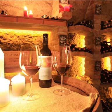 A cozy wine cellar with candlelight. On a wooden barrel are two wine glasses and a bottle of red wine.