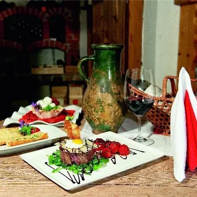 A beautifully arranged table with a dish of meat, fresh vegetables, and a side dish. Next to it are a carafe, a glass of red wine, and napkins.