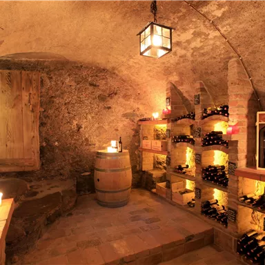 A cozy wine room with wooden shelves filled with bottles. Soft light from candles and a lamp creates an inviting atmosphere.