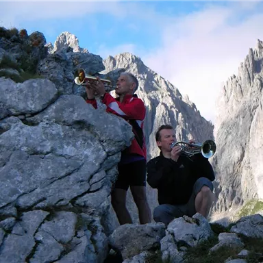 Zwei Männer spielen Trompete auf einem Felsen in den Bergen. Im Hintergrund sind beeindruckende Berggipfel und ein klarer Himmel zu sehen.