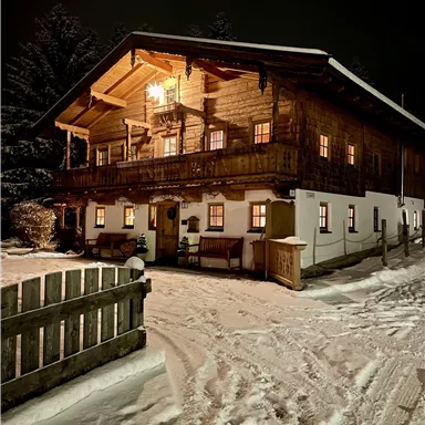 Ein gemütliches Holzhaus im Schnee bei Nacht, beleuchtet von warmem Licht. Der Weg zum Haus ist verschneit und bietet eine einladende Atmosphäre.