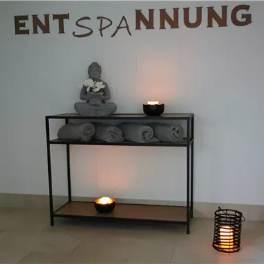 Ein ruhiger Raum mit einer Buddha-Statue und gefalteten Handtüchern. Sanftes Licht von Kerzen schafft eine entspannende Atmosphäre.