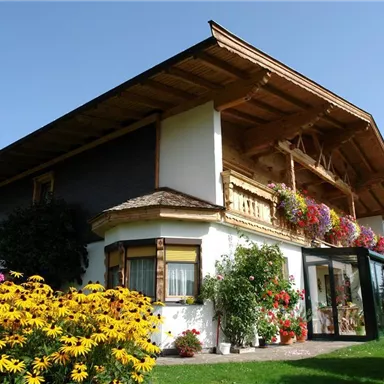 Ein charmantes Haus mit einem Holzbalkon und bunten Blumen. Der Garten ist üppig und grün, umgeben von Bergen im Hintergrund.