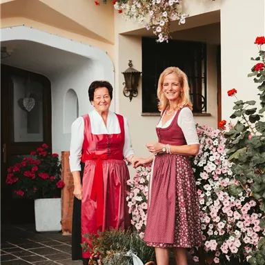 Two women in traditional costumes stand in front of a blooming plant arrangement. The entrance area is inviting and colorful.