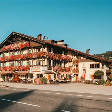 A picturesque hotel with blooming balconies and traditional architecture. In the background, gentle hills and a blue sky can be seen.