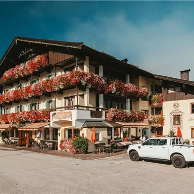 A charming alpine hotel with blooming flower balconies. In front of the building, there is a white car parked in the parking lot.
