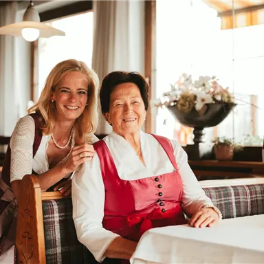Two women smile in a cozy restaurant. The older lady is wearing a traditional outfit, while the younger woman is posing in elegant clothing.