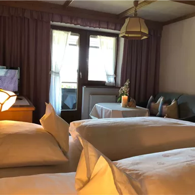 A cozy room with two beds and a small table. Windows with curtains and a seating area create an inviting atmosphere.