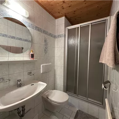 A modern bathroom with a shower, a sink, and a round mirror. The walls are tiled and there is a wooden ceiling covering.