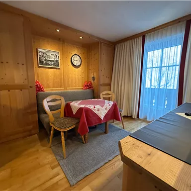 A cozy room with wooden walls and a small dining area. A table with a red tablecloth and a window with curtains create an inviting atmosphere.