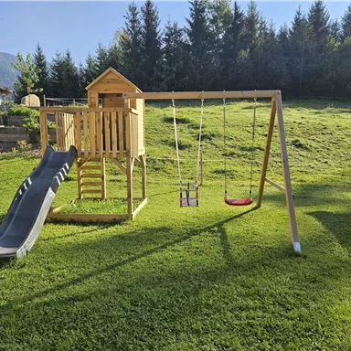 A playground with a slide and two swings is situated in a green garden. In the background, trees and a light blue sky are visible.