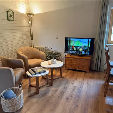 A cozy living room with two beige armchairs, a table, and a television. The room has wooden floors and large windows with curtains.