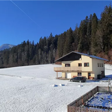 Ein gemütliches Haus steht auf einem schneebedeckten Feld, umgeben von Bäumen. Im Hintergrund sind Berge und ein klarer blauer Himmel sichtbar.