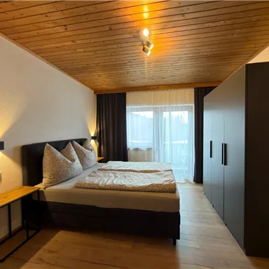 Ein modernes Schlafzimmer mit einem großen Bett und gemütlichen Kissen. Große Fenster lassen viel Tageslicht herein.