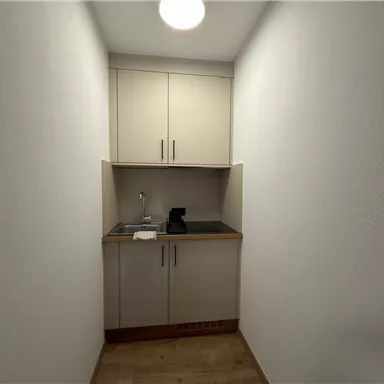 Eine kleine Küchenzeile in einem schmalen Raum. Sie enthält einen Wasserhahn, eine Kaffeemaschine und moderne Schränke.