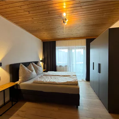 A modern bedroom with a large bed and wooden ceiling. The room has large windows and a stylish wardrobe.