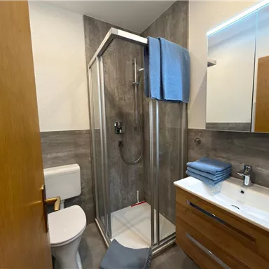 A modern bathroom with a shower and wooden shelves. The walls are covered with gray tiles, and there are towels and toiletries on the sink.