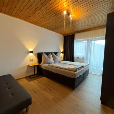 A cozy bedroom with a double bed and two pillows. There is a sofa and large windows that let in plenty of daylight.