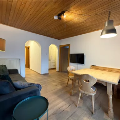 A cozy living room with a sofa, a table, and chairs. The room has a bright wooden ceiling and a television on the wall.