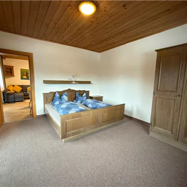 A cozy bedroom with a double bed and blue pillows. In the background, there is a wardrobe and an open door to another room.