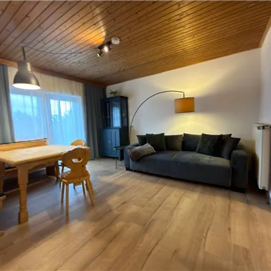 A cozy living room with a wooden table, a couch, and large windows. The decor is simple and modern, with warm wood and soft colors.