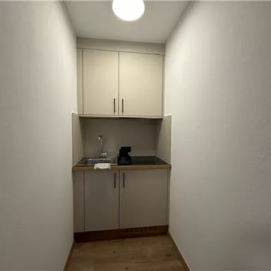 A small kitchen with a sink and a coffee machine. The walls are white and the floor is wooden.