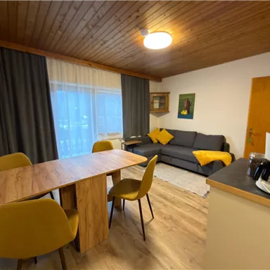 A cozy living space with a dining table and yellow chairs. In the background, there is a couch and a large window front that lets in natural light.