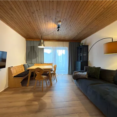 A modern living room with a wooden ceiling, a cozy sofa, and a dining area. Large windows let in plenty of daylight.