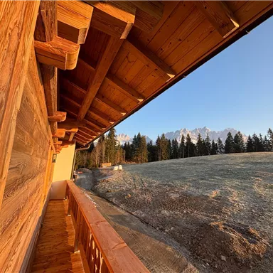 A wooden porch overlooking a picturesque mountain landscape. In the background, snow-covered peaks and dense forests can be seen.