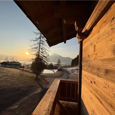 A view from a balcony of a picturesque landscape at sunrise. In the background, mountains and trees can be seen, while the sun illuminates the sky.