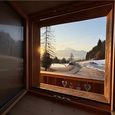 A beautiful view from a window overlooking a snow-covered landscape with mountains in the background. The sun is rising, casting warm light over the scene.