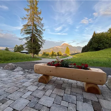 A picturesque landscape with a wooden table and flowers in the foreground. In the background, majestic mountains rise under a clear sky.