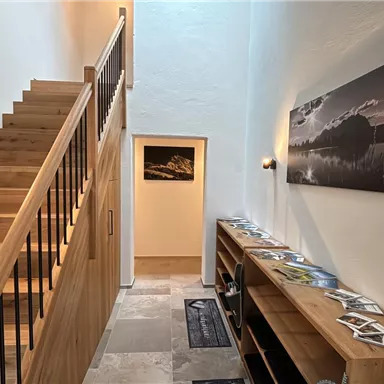 A modern entrance area with a wooden staircase and bright walls. A large photo hangs on the wall, and brochures are lying on a shelf.