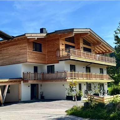 A modern wooden house with several balconies and green surroundings. The sun is shining under a blue sky on the building.