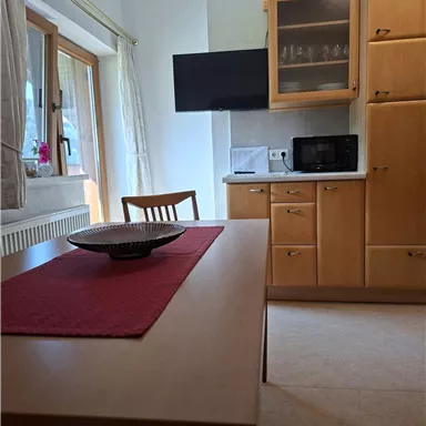 A cozy kitchen with a wooden table and a television. The furniture is made of light wood and there is a microwave as well as windows with daylight.