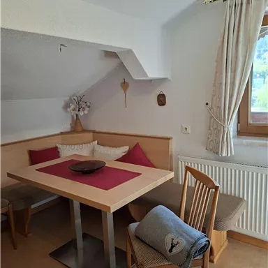A cozy dining area with a table and chairs. The walls are bright, and there are windows with curtains that let in a lot of light.
