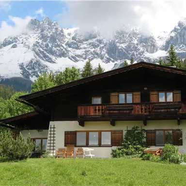 A pretty alpine house with a wooden balcony, surrounded by green meadows. In the background, majestic snow-covered mountains rise under a clear sky.