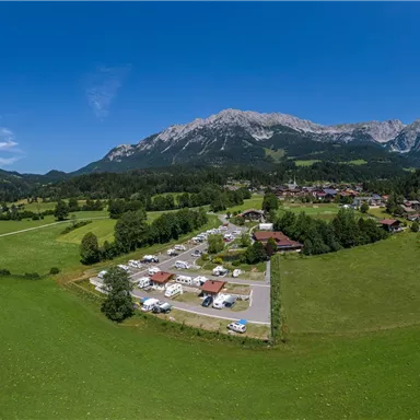 Eine weite, grüne Landschaft mit Bergen im Hintergrund. In der Mitte sind Wohnmobile und ein kleiner Ort sichtbar.
