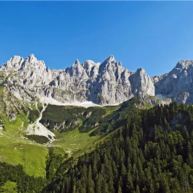 An impressive mountain landscape with high peaks and lush green meadows. The sky is clear and blue.