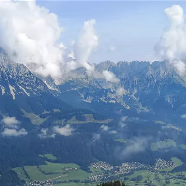 Eine beeindruckende Berglandschaft mit hohen Gipfeln und weißen Wolken. Im Vordergrund sind grüne Wiesen und kleine Siedlungen zu sehen.