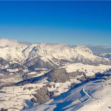 An impressive winter landscape with snow-covered mountains and a clear blue sky. The view shows wide snowy valleys and gentle hills.