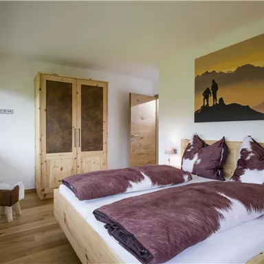 Ein gemütliches Schlafzimmer mit einem großen Bett und Kissen in Braun- und Weißtönen. An der Wand hängt ein Bild mit einer Berglandschaft und Silhouetten von Wanderern.