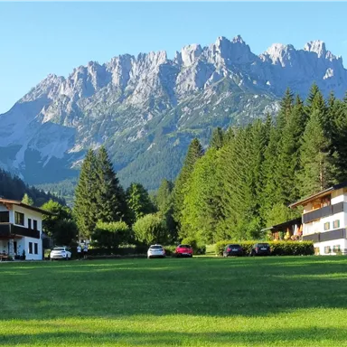 A picturesque landscape with two buildings and a large, green lawn. In the background, an impressive mountain range rises.