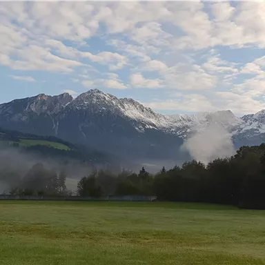 Eine malerische Berglandschaft mit schneebedeckten Gipfeln und sanften Wolken. Im Vordergrund erstreckt sich eine grüne Wiese.