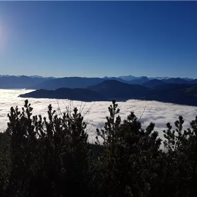 Eine beeindruckende Berglandschaft mit Wolken, die wie ein Meer unterhalb wirken. Der Himmel ist klar und sonnig.
