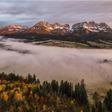 Eine malerische Landschaft mit Bergen und einer Nebeldecke über den Wiesen. Bunte Bäume im Vordergrund verleihen der Szene eine herbstliche Note.