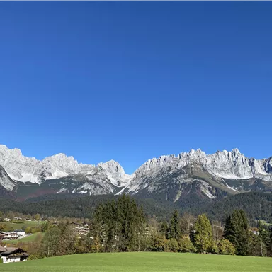 Eine beeindruckende Berglandschaft mit schneebedeckten Gipfeln und einem klaren blauen Himmel. Im Vordergrund sind grüne Wiesen und einige Häuser zu sehen.